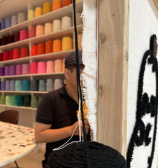 Black yarn is attached to a frame to allow the outline of a rug to be tufted onto a framed fabric. A range of colorful yarns are placed in the background, awaiting to form part of the art piece.