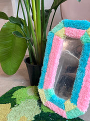 A mirror in a tufted frame with pastel colours placed next to a plant.