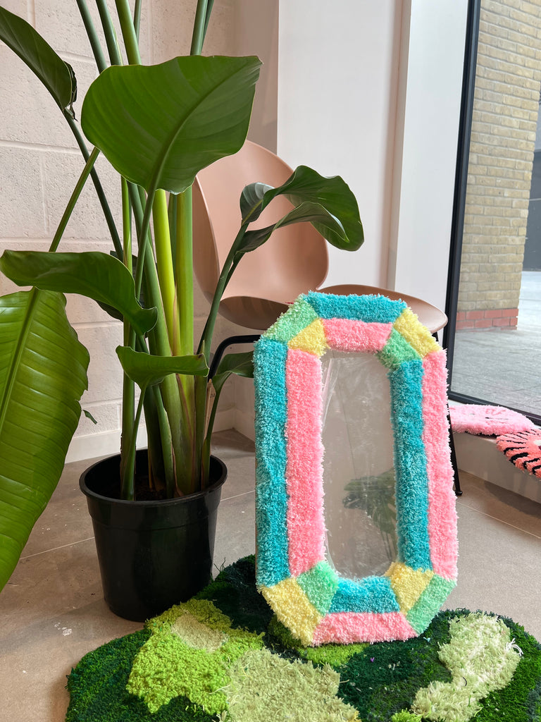 A multi-coloured tutfted mirror frame rests on the leg of a chair, on top of another green textured rug.