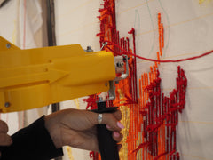 Tufting gun on a fabric canvas creating red and orange lines, forming part of an art piece.